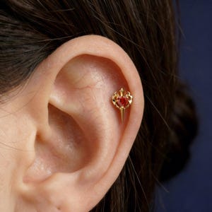 May include: A gold-colored ear piercing with a red gemstone center. The piercing has a crown-like design at the top and a pointed bottom. The jewelry is set against the backdrop of a person's ear.