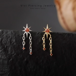 Pode incluir: Brincos com design de estrela, com uma pedra vermelha central e detalhes de corrente pendurada. Um brinco é prateado, o outro dourado. São joias corporais, conforme indicado no texto "Vivi Piercing Jewelry".