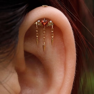 Könnte beinhalten: Ein goldfarbenes Ohrpiercing mit einem herzförmigen roten Edelstein, akzentuiert mit kleinen weißen Edelsteinen und hängenden Ketten, die in spitzen Formen enden. Ein kleiner, runder goldener Ohrstecker ist ebenfalls sichtbar.