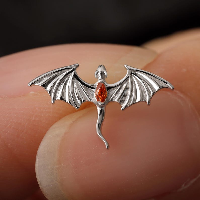 May include: A silver dragon pendant with outstretched wings and an orange gemstone set in the center. The dragon has detailed wing and body features. The pendant is a jewelry item.