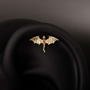 May include: A gold-colored dragon-shaped earring with a red gemstone in the center. The dragon has outstretched wings and a long tail. The earring is a small, detailed piece of jewelry.