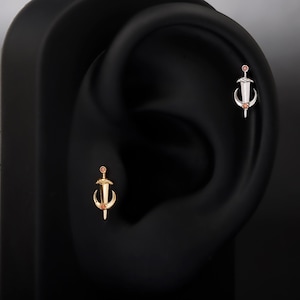 Puede incluir: Primer plano de dos piercings en la oreja. Uno es dorado, el otro plateado. Ambos presentan un diseño de espada con una pequeña gema redonda de color naranja-rojizo. Los pendientes están colocados sobre un fondo negro, resaltando los detalles de la joyería.