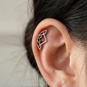 Könnte beinhalten: Ein silberfarbenes Knorpel-Piercing für das Ohr in einer geometrischen, fächerartigen Form mit schwarzer Emaille-Einlage. Das Design besteht aus einem geschwungenen Außenrahmen mit mehreren innenliegenden, blütenblattartigen Segmenten.