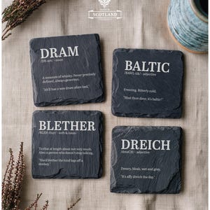 May include: Four square dark grey slate coasters with rough, natural edges. Each coaster features a white printed dictionary-style definition for the Scottish words Dram, Baltic, Blether, and Dreich, including phonetic pronunciations and example sentences.