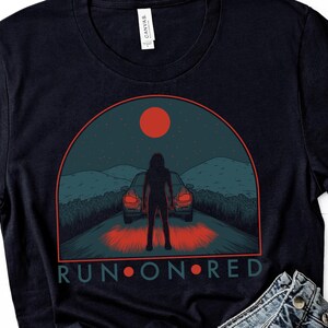 May include: Black t-shirt with a graphic of a person standing in front of two cars with red taillights. The graphic is set against a dark blue background with a red moon and the text "RUN ON RED".