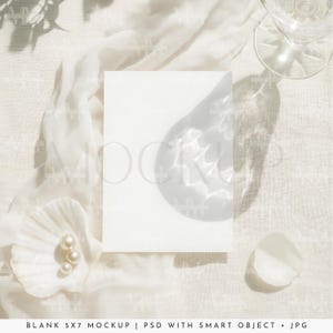 Puede incluir: Una maqueta de tarjeta blanca de 5x7 pulgadas con una suave superposición de sombras, sobre una tela blanca texturizada. Una concha con perlas, una copa de vino y un pétalo están dispuestos alrededor de la tarjeta, creando una estética minimalista.
