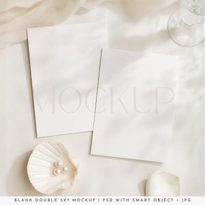 Puede incluir: Maqueta con dos tarjetas blancas en blanco de 5x7 pulgadas, una concha con perlas y un vaso sobre una superficie blanca. La palabra "MOCKUP" es visible en las tarjetas. La escena está suavemente iluminada.