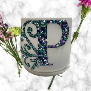 May include: White ceramic mug with a large, jeweled letter "P" in shades of teal, purple, and green. The letter is embellished with sparkling rhinestones. A decorative swirl design in teal rhinestones is on the left side of the mug. Pink and purple flowers are in the background.