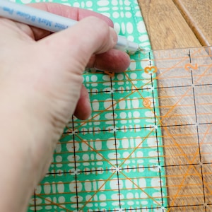 May include: A hand using a ruler and a pen to mark a piece of fabric with a turquoise and white pattern.