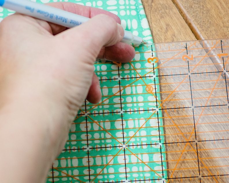 May include: A hand using a ruler and a pen to mark a piece of fabric with a turquoise and white pattern.