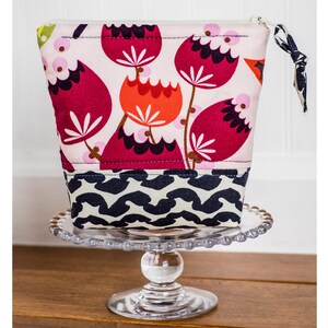 May include: A pink and red floral print zippered pouch with a black and white geometric print bottom. The pouch is sitting on a clear glass cake stand.