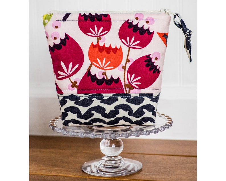May include: A pink and red floral print zippered pouch with a black and white geometric print bottom. The pouch is sitting on a clear glass cake stand.
