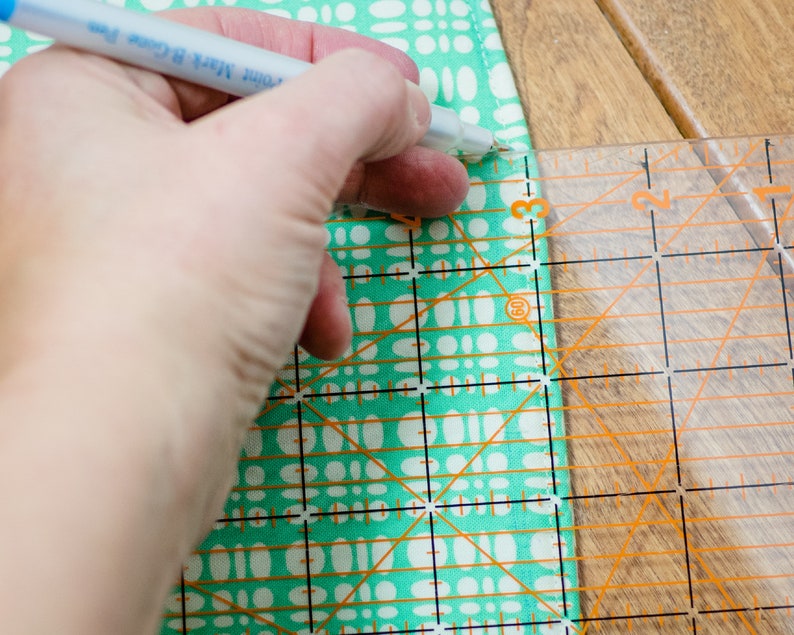 K&ouml;nnte beinhalten: Eine Hand, die mit einem Lineal und einem Stift ein St&uuml;ck Stoff mit einem t&uuml;rkisfarbenen und wei&szlig;en Muster markiert.