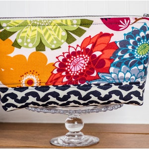 May include: A colorful floral zippered pouch with a black and white geometric pattern on the bottom. The pouch is sitting on a clear glass pedestal.