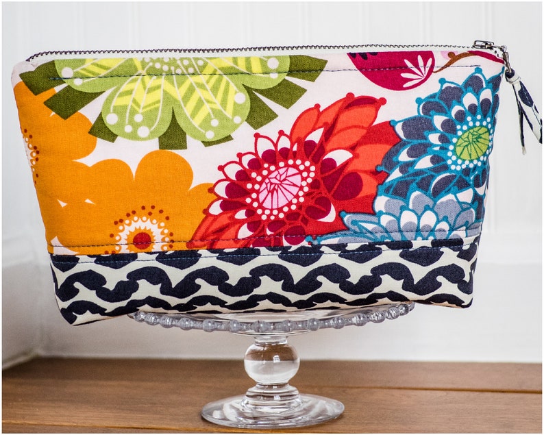 May include: A colorful floral zippered pouch with a black and white geometric pattern on the bottom. The pouch is sitting on a clear glass pedestal.