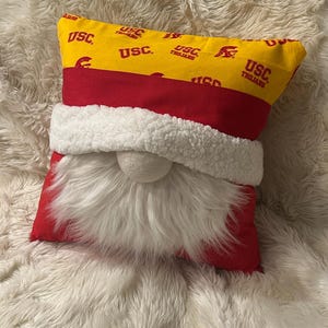 May include: A decorative pillow in the shape of a gnome. The top is yellow with the text "USC Trojans" in red. The middle is red, and the bottom is red with a white beard and a white ball nose. The pillow is on a white fluffy surface.