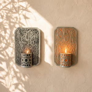 May include: Two rectangular wall sconces with textured backplates. The left sconce features a dark gray, rough-textured backplate and a cylindrical candle holder with a geometric cutout pattern. The right sconce has a light brown, wavy-textured backplate and a matching cylindrical candle holder. Both hold lit candles.