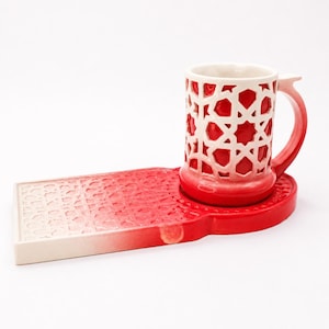 Può includere: Un set composto da una tazza in ceramica e un vassoio rettangolare abbinato, caratterizzato da una sfumatura di colore rosso e crema. Entrambi i pezzi sono decorati con un motivo geometrico in rilievo che rivela la base rossa sottostante.