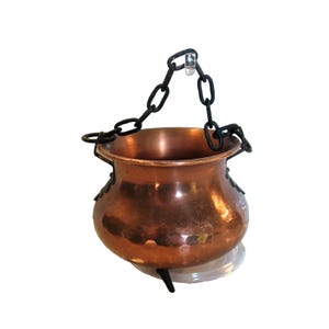 May include: A polished copper pot with a rounded body and a wide opening. The pot features three small black metal legs at the base and a black metal chain handle attached to two side rings, allowing it to be suspended from a hook.