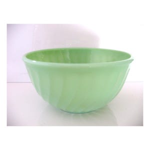 May include: A light green, vintage glass mixing bowl. The bowl has a rounded shape with a wide opening and a series of vertical, curved ridges on the exterior. The bowl is made of a solid, opaque glass.