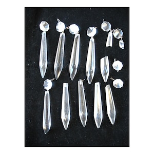 May include: A collection of clear crystal chandelier prisms and decorative faceted crystal beads. The prisms are elongated, with a pointed bottom and a faceted top. The beads are round and multi-faceted. The crystals are displayed on a black background.