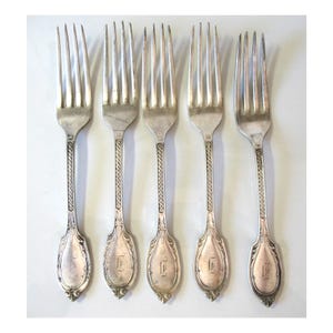 May include: A set of five silver-toned metal forks arranged in a row. Each fork features four tines, a slender handle with a repeating rope-like embossed pattern, and a flared base engraved with a stylized letter E in a cursive script.