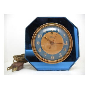 May include: A vintage electric clock with a blue mirrored octagonal frame. The clock face is blue with gold numerals and hands, and a gold-colored center. The clock has a brown cord and plug.