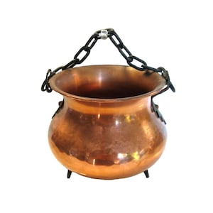May include: A round copper pot with a polished finish, featuring three small feet at the base and a black metal chain handle attached to two side mounts. The vessel has a wide, open top and a curved, bulbous body shape.