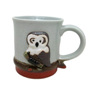 May include: A ceramic mug with a light gray speckled finish. The mug features a 3D owl design in brown, white, and black. The base of the mug is red, and the handle is light gray. A unique and decorative item for coffee or tea.
