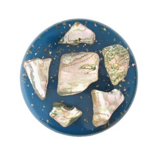 May include: A round, blue decorative object with iridescent abalone shell pieces. The shell pieces are various shapes and sizes, and are arranged on the blue surface. Small, gold-colored flecks are scattered around the shell pieces.