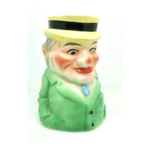 May include: A vintage ceramic figurine of a man wearing a yellow hat with a black band and a green jacket. The figurine has a smiling face with rosy cheeks, blue eyes, and a red mouth. A blue tie is visible.