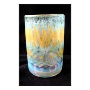 May include: A cylindrical glass with a colorful, swirled design. The glass features bands of yellow and light blue, with a darker blue base. The rim has a textured appearance, and the glass is set against a black background.
