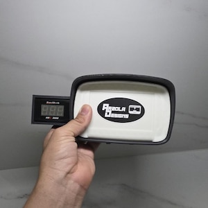 May include: A black and white rectangular device with the text "FuelTech" and a digital display showing "888". The device is attached to a white rectangular box with the text "Arzola Designs" in black.