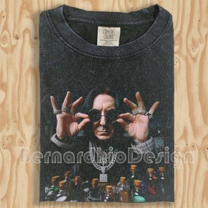 May include: Dark gray Comfort Colors t-shirt featuring a portrait of a man wearing sunglasses and jewelry. The image includes bottles and the text "BernardbioDesign". The shirt has a crew neck and a tag.