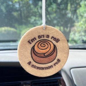 May include: A round, tan car air freshener with the text "I'm on a roll" and "A cinnamon roll" surrounding a graphic of a cinnamon roll. The air freshener hangs from a white string.