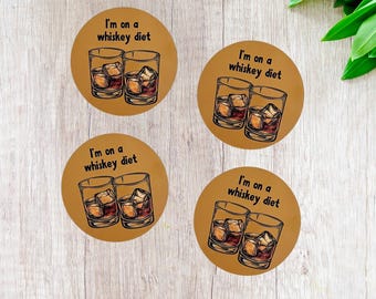 May include: Four round brown coasters with the text "I'm on a whiskey diet" and a graphic of two whiskey glasses with ice cubes. The coasters are arranged on a light brown wooden surface.