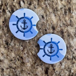 May include: Two round car coasters with a nautical theme. Each coaster features a blue and white camouflage pattern and a navy blue ship's wheel design with an anchor in the center.