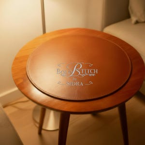 May include: A circular, brown leather placemat on a small, wooden side table. The placemat is a lighter shade of brown than the table. The table has three legs and a dark wood finish. The text "Bold Stitch by Sidra" is printed on the placemat.