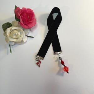 May include: Black velvet choker necklace with silver-toned metal ends. The choker features a red heart charm and a cluster of red beads. A pink and white rose floral arrangement is also in the image.