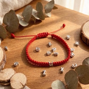 May include: A red braided bracelet with silver letter beads spelling out a name or word. The bracelet is on a wooden surface with scattered silver letter beads and decorative leaves.