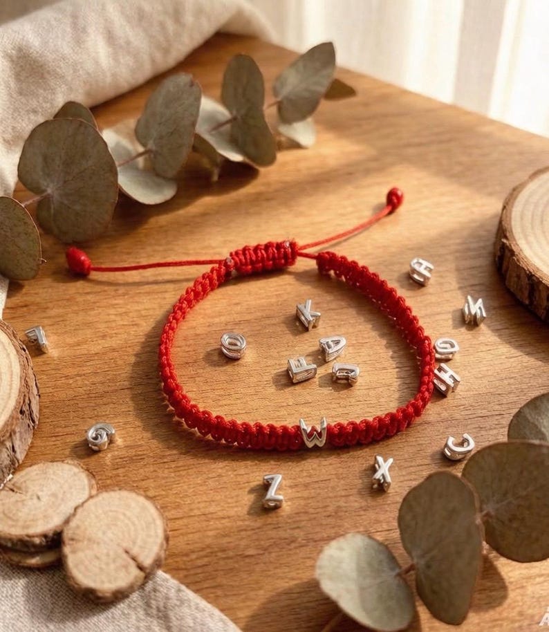 May include: A red braided bracelet with silver letter beads spelling out a name or word. The bracelet is on a wooden surface with scattered silver letter beads and decorative leaves.