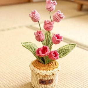May include: A handcrafted crochet flower arrangement featuring pink tulip blooms, green leaves, and stems, presented in a beige and yellow pot. The pot has a small brown tag with text and a smiling face.