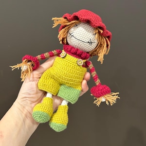 May include: A handmade crochet doll resembling a scarecrow. The doll features a white head with stitched facial features, a red hat, and straw-like hair. It wears yellow overalls with green accents and red and brown striped arms.