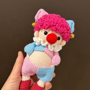 May include: A handmade crochet clown doll with a pink, blue, and beige color scheme. The clown has a red nose, a pink curly wig, and a white ruffled collar. The doll is wearing a blue and pink outfit and red shoes.