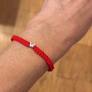 May include: A red woven bracelet with a silver letter "W" charm. The bracelet is made of a thick, braided cord and is worn on a wrist. The background is a blurred wooden surface.