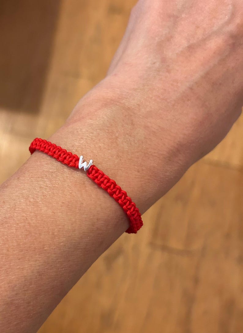 May include: A red woven bracelet with a silver letter "W" charm. The bracelet is made of a thick, braided cord and is worn on a wrist. The background is a blurred wooden surface.