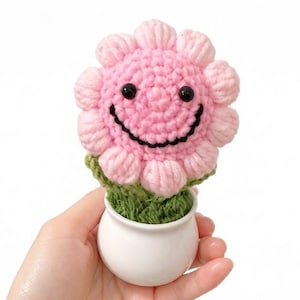 May include: A handmade crochet flower with a smiling face, in a small white ceramic pot. The flower is pink with black eyes and a black smile. The pot is filled with green yarn, resembling grass.