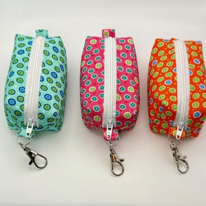 May include: Three small, rectangular fabric pouches with white zippers and silver-colored clasps. The pouches are patterned with colorful floral designs on a teal, pink, and orange background. The pouches are likely used for carrying small items.