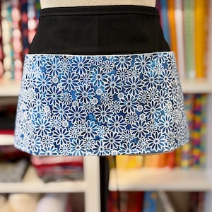 May include: A black waist apron with a blue floral pattern. The apron features a solid black waistband and a skirt adorned with white daisy-like flowers on a blue background. The apron is displayed on a mannequin.