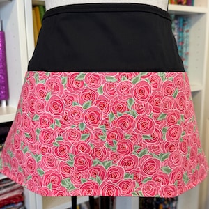May include: A black waist apron with a vibrant pink rose pattern. The apron features a solid black waistband and a skirt adorned with a repeating design of pink roses and green leaves. This apron is suitable for kitchen or craft use.